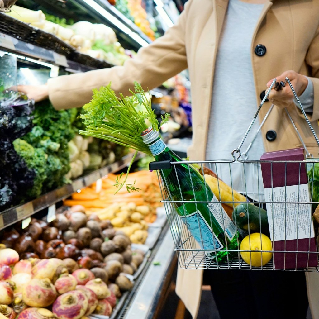 Woman in a white coat pushing a green shopping cart through a grocery store produce section