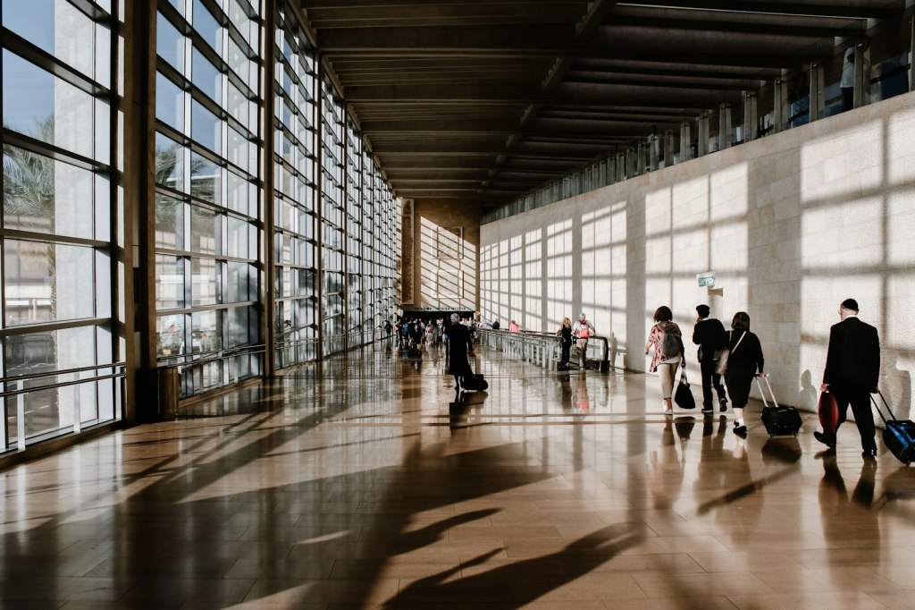 Travelers walking through a sunlit airport terminal with large glass windows