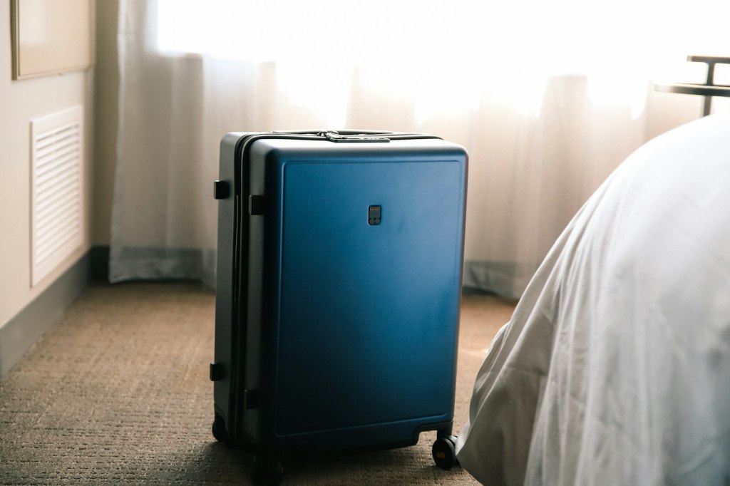 A blue suitcase sitting on the floor next to a made hotel bed