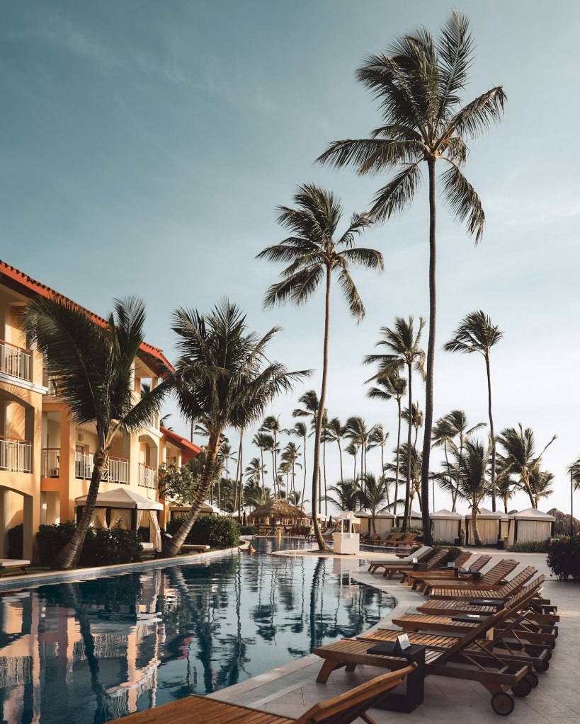 Wooden lounge chairs beside a resort pool surrounded by palm trees
